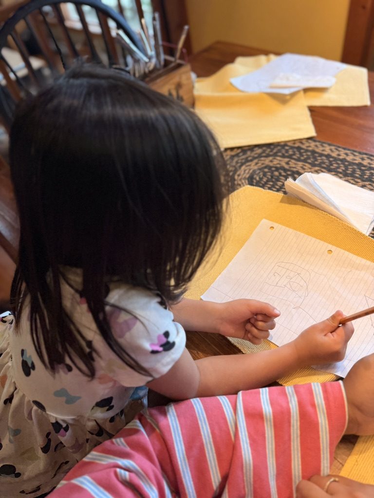Olive, seen from behind, drawing a birdhouse design at the kitchen table