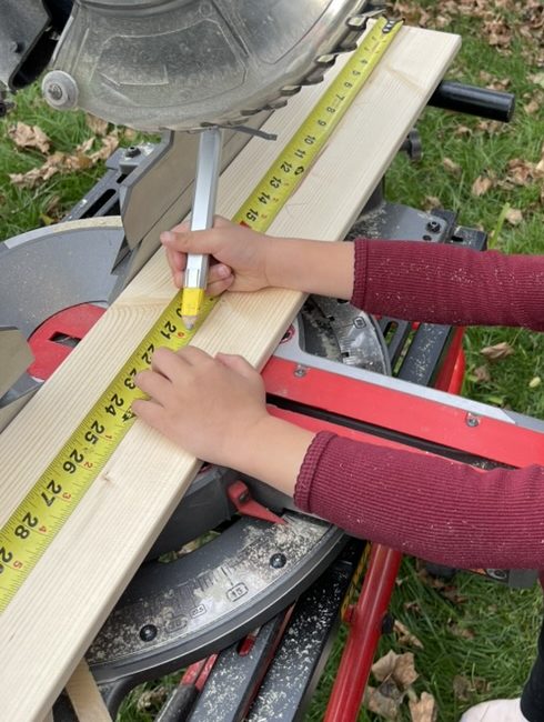 Olive's hands marking a board with a carpenter's pencil along a yellow tape measure