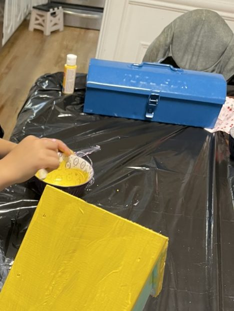A hand dipping a brush into a bowl of yellow paint next to the birdhouse's freshly painted yellow roof