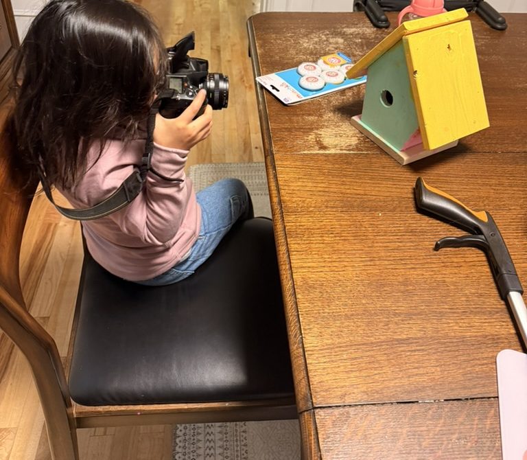 Olive, seen from behind, photographing the birdhouse with a big camera